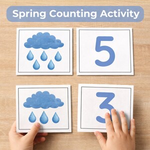 May include: A spring counting activity set with cards featuring blue clouds, raindrops, and numbers 3 and 5. The text "Spring Counting Activity" is displayed at the top. The cards are on a light brown wooden surface.
