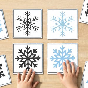 Può includere: Diverse carte quadrate con disegni di fiocchi di neve neri e azzurri sono disposte su una superficie di legno chiaro. Le mani di un bambino interagiscono con alcune delle carte. I fiocchi di neve presentano motivi simmetrici dettagliati.