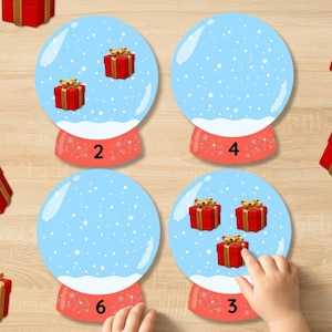 May include: An educational game with snow globes, each containing a different number of red gift boxes. The globes are on a light wooden surface, with more gifts scattered around. A child's hand is interacting with the game.
