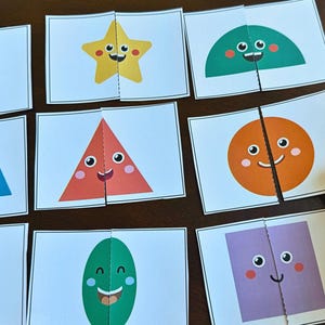 May include: A set of colourful, two-piece shape puzzles featuring smiling faces. Shapes include a heart, star, semi-circle, triangle, circle, trapezoid, oval, and square. The puzzles are on white cards.