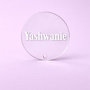 Puede incluir: Un objeto circular transparente de acrílico con el nombre "Yashwanie" en blanco, con fuente sans-serif. Tiene un pequeño agujero en la parte inferior y está sobre un fondo morado claro. El diseño es simple y moderno.