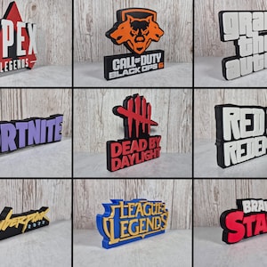 Könnte beinhalten: Eine Sammlung farbenfroher 3D-Schilder mit beliebten Videospieltiteln. Die Schilder zeigen "Apex Legends", "Call of Duty Black Ops 6", "Grand Theft Auto", "Fortnite", "Dead by Daylight", "Red Dead Redemption", "Cyberpunk", "League of Legends" und "Brawl Stars".