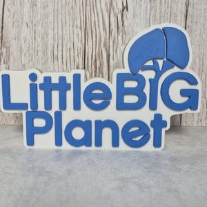 May include: A blue and white sign displaying the text "Little BIG Planet". The sign has a blue graphic above the word "BIG". The sign is set against a grey surface with a wooden backdrop.