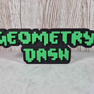 May include: A black and green "GEOMETRY DASH" sign. The text is in a pixelated font, with green letters on a black background. The sign is likely a decorative item or a piece of merchandise related to the game.