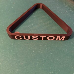 May include: A black triangle-shaped pool ball rack with the word "CUSTOM" printed in white letters.