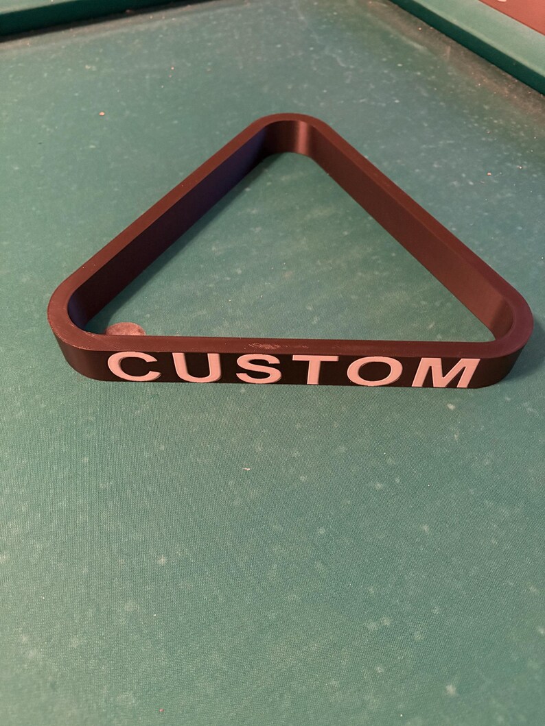 May include: A black triangle-shaped pool ball rack with the word "CUSTOM" printed in white letters.