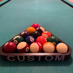 May include: A black triangle-shaped pool ball rack with the word "CUSTOM" printed on the front. The rack is filled with 15 billiard balls, including a red ball, a yellow ball, a blue ball, a green ball, and a black ball.