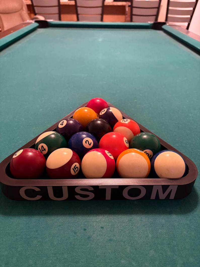 May include: A black triangle-shaped pool ball rack with the word "CUSTOM" printed on the front. The rack is filled with 15 billiard balls, including a red ball, a yellow ball, a blue ball, a green ball, and a black ball.