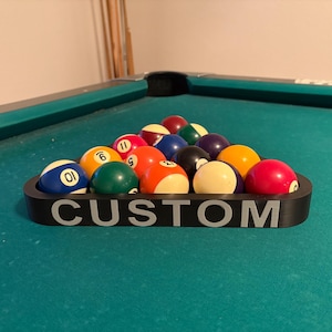 May include: A black triangle-shaped pool ball rack with the word "CUSTOM" printed on the front. The rack is filled with 15 billiard balls, including the cue ball, numbered balls, and striped balls.