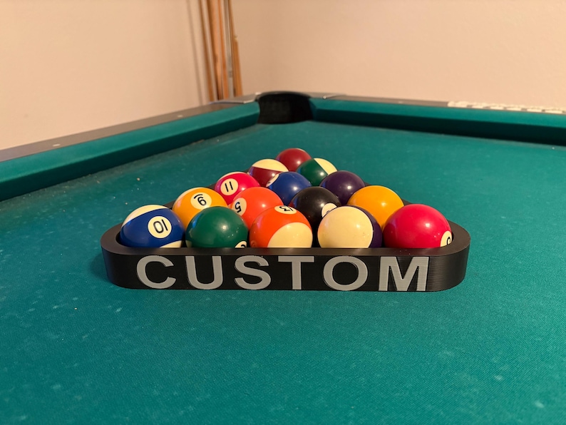 May include: A black triangle-shaped pool ball rack with the word "CUSTOM" printed on the front. The rack is filled with 15 billiard balls, including the cue ball, numbered balls, and striped balls.