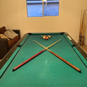 May include: A green felt pool table with two crossed cues and a set of billiard balls arranged in a triangle on the table. The table has a black felt edge and a black and silver base.