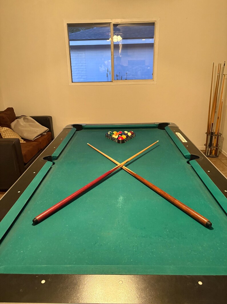 May include: A green felt pool table with two crossed cues and a set of billiard balls arranged in a triangle on the table. The table has a black felt edge and a black and silver base.