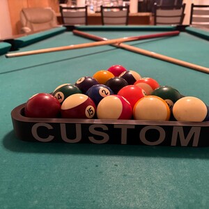 May include: A black pool ball rack with the word "CUSTOM" printed on the front. The rack is filled with 15 billiard balls, including a solid red ball, a solid yellow ball, a solid blue ball, a solid green ball, a solid purple ball, a solid orange ball, a solid brown ball, a solid black ball, a striped red ball, a striped yellow ball, a striped blue ball, a striped green ball, a striped purple ball, a striped orange ball, and a striped brown ball.