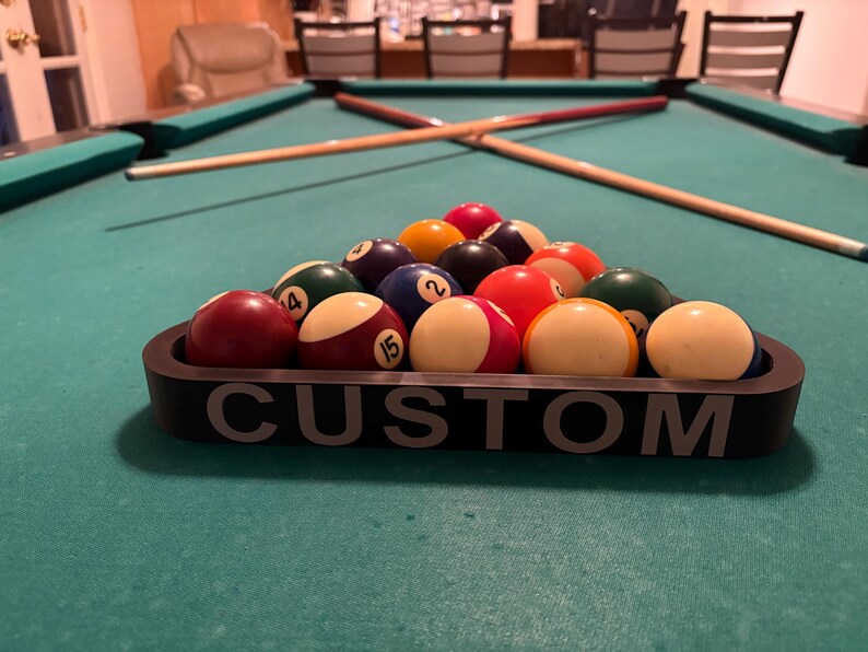 May include: A black pool ball rack with the word "CUSTOM" printed on the front. The rack is filled with 15 billiard balls, including a solid red ball, a solid yellow ball, a solid blue ball, a solid green ball, a solid purple ball, a solid orange ball, a solid brown ball, a solid black ball, a striped red ball, a striped yellow ball, a striped blue ball, a striped green ball, a striped purple ball, a striped orange ball, and a striped brown ball.