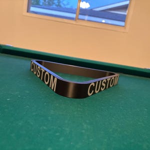 May include: A black pool table rack with the word "CUSTOM" printed on both sides in white lettering.