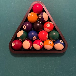 May include: A black triangle-shaped rack holds a set of 15 billiard balls, including the cue ball. The balls are arranged in a pyramid shape, with the cue ball at the top. The balls are numbered 1 through 15, and each ball has a different color.