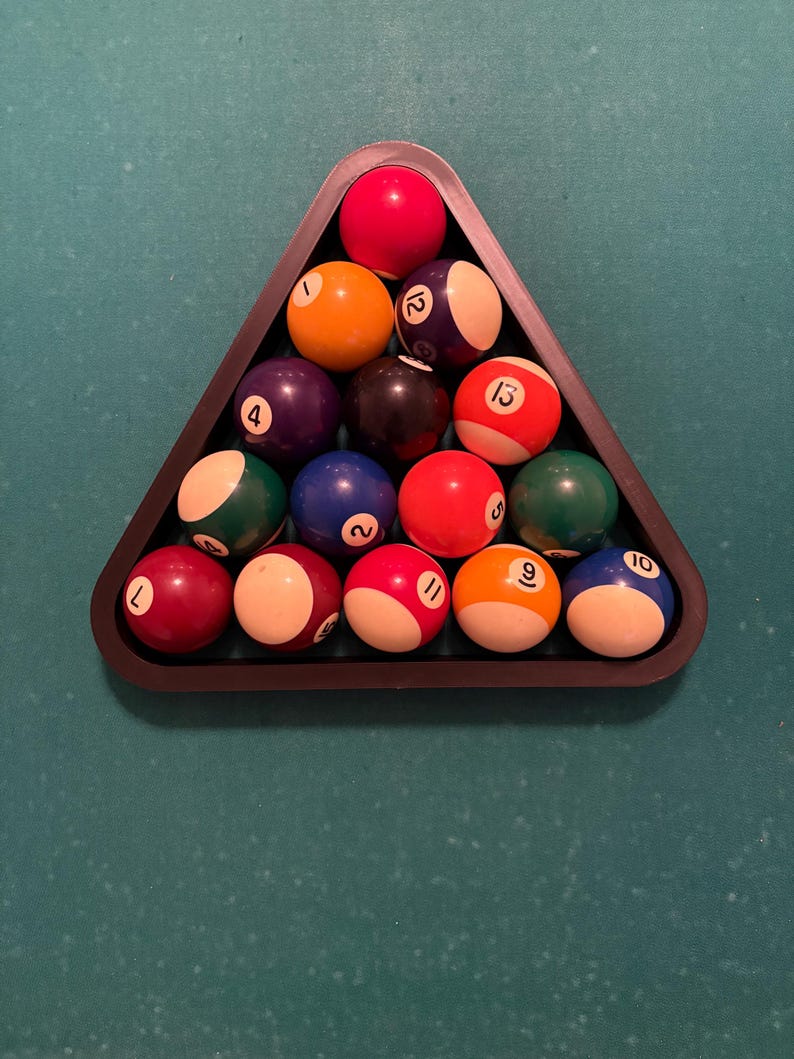 May include: A black triangle-shaped rack holds a set of 15 billiard balls, including the cue ball. The balls are arranged in a pyramid shape, with the cue ball at the top. The balls are numbered 1 through 15, and each ball has a different color.