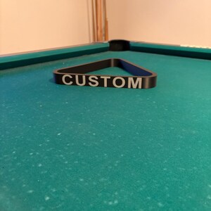 May include: A black pool triangle rack with the word "CUSTOM" printed in white on the front.
