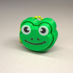 May include: A green frog-shaped keyring with a smiling face, white eyes, and black pupils. The keyring has a yellow ball chain for attachment. The frog's design is simple and cartoonish.