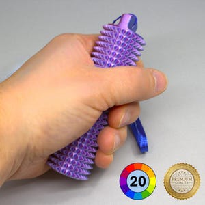 May include: A purple, spiked massage tool with a blue lanyard is held in a hand. The tool has a series of triangular spikes. A colour wheel with the number 20 and a gold "Premium Quality" seal are in the lower right corner.