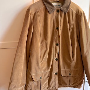 May include: A tan corduroy jacket featuring a dark brown collar, a front button closure, and two lower front pockets with buttoned flaps. The interior label displays the text L.L.Bean. The jacket is displayed on a wooden hanger.