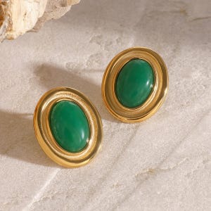 Könnte beinhalten: Ein Paar goldfarbene Ohrstecker mit ovalen, smaragdgrünen Mittelstücken. Die Ohrringe haben einen doppelwandigen, kreisförmigen Goldrahmen. Die Ohrringe werden auf einer hellen, strukturierten Oberfläche präsentiert.