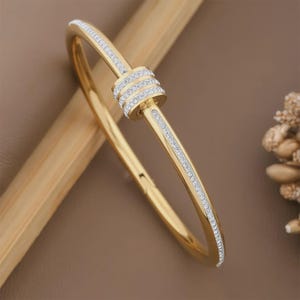 May include: Gold-toned bangle bracelet with a central cylindrical design and sparkling clear stones. The bracelet features a row of stones along the outer edge. The jewelry is displayed on a brown surface.