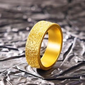 Puede incluir: Un anillo de oro texturizado con una banda ancha. El anillo tiene una apariencia rugosa y martillada, que captura la luz y crea un efecto brillante. Es una joya simple pero elegante.