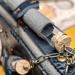May include: A close-up of a small glass vial with a cork stopper, attached to a brass chain. The vial is filled with a light-colored substance. The vial is attached to a gray fabric and dark blue leather case.