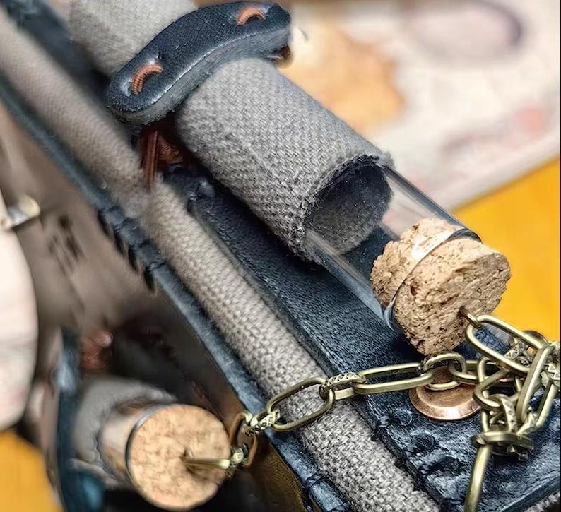 May include: A close-up of a small glass vial with a cork stopper, attached to a brass chain. The vial is filled with a light-colored substance. The vial is attached to a gray fabric and dark blue leather case.