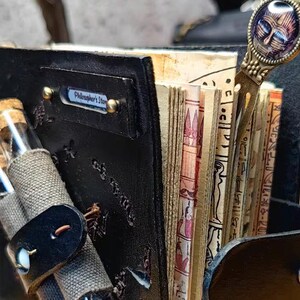 May include: A black leather journal with the text "Philosopher's Story" on a metal plate. The journal is open, revealing aged pages with illustrations and hieroglyphs. A decorative bookmark and small glass vials are also visible.