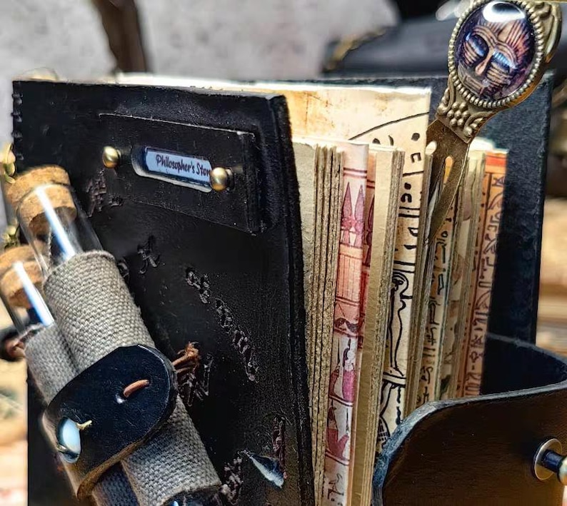 May include: A black leather journal with the text "Philosopher's Story" on a metal plate. The journal is open, revealing aged pages with illustrations and hieroglyphs. A decorative bookmark and small glass vials are also visible.