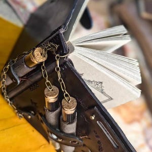 May include: A black leather journal with a decorative chain and two small glass vials with cork stoppers. The journal's pages are visible, and the cover has a detailed design. The vials are suspended by a gold-colored chain.