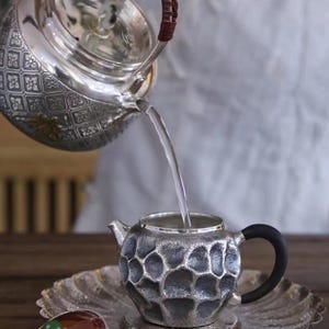Puede incluir: Una tetera plateada vierte agua en una peque&ntilde;a taza de t&eacute; plateada texturizada con un asa negra. La tetera tiene un dise&ntilde;o estampado y la taza de t&eacute; se encuentra sobre un platillo a juego. Una tapa con un bot&oacute;n verde descansa cerca.