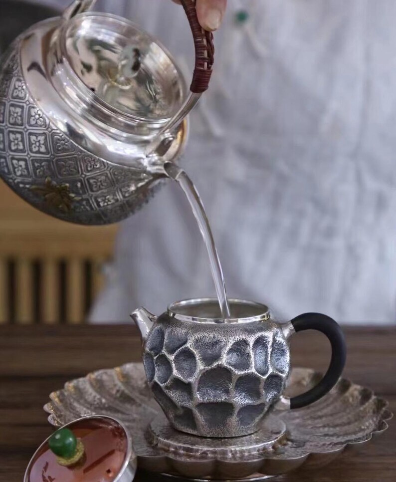 Puede incluir: Una tetera plateada vierte agua en una peque&ntilde;a taza de t&eacute; plateada texturizada con un asa negra. La tetera tiene un dise&ntilde;o estampado y la taza de t&eacute; se encuentra sobre un platillo a juego. Una tapa con un bot&oacute;n verde descansa cerca.