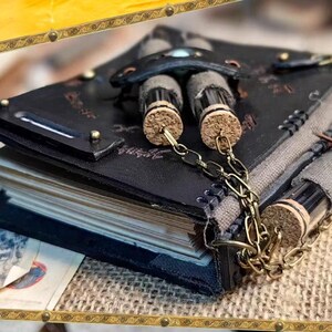 May include: A handmade, black leather journal with a unique design. The journal features a decorative clasp, brass accents, and a chain holding small glass vials with cork stoppers. The pages are visible, suggesting a blank notebook.