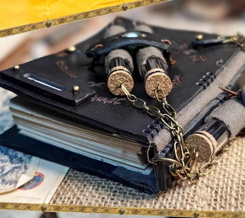 May include: A handmade, black leather journal with a unique design. The journal features a decorative clasp, brass accents, and a chain holding small glass vials with cork stoppers. The pages are visible, suggesting a blank notebook.