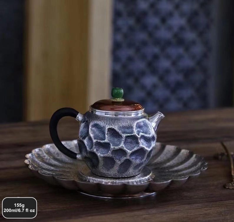 Puede incluir: Una tetera plateada con asa negra y tapa marr&oacute;n, sobre un platillo a juego. La tetera tiene un patr&oacute;n texturizado en forma de panal. La tapa est&aacute; rematada con una gema verde. Contiene 200ml y pesa 155g.