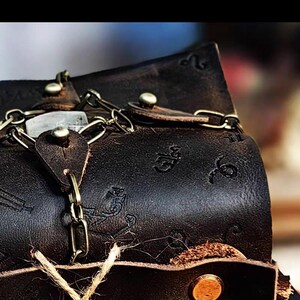 May include: Close-up of a dark brown leather bag with a chain and clasp closure. The bag has embossed designs and metal rivets. A natural twine is attached to the bag.