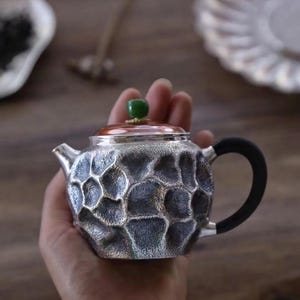 Puede incluir: Peque&ntilde;a tetera plateada con un patr&oacute;n texturizado en forma de panal. Cuenta con un asa negra, un borde color cobre y un bot&oacute;n verde en la tapa. La tetera se sostiene en una mano, lo que sugiere su tama&ntilde;o compacto y est&aacute; dise&ntilde;ada para t&eacute;.