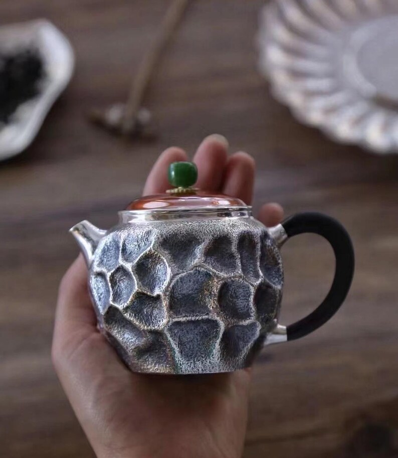 Puede incluir: Peque&ntilde;a tetera plateada con un patr&oacute;n texturizado en forma de panal. Cuenta con un asa negra, un borde color cobre y un bot&oacute;n verde en la tapa. La tetera se sostiene en una mano, lo que sugiere su tama&ntilde;o compacto y est&aacute; dise&ntilde;ada para t&eacute;.