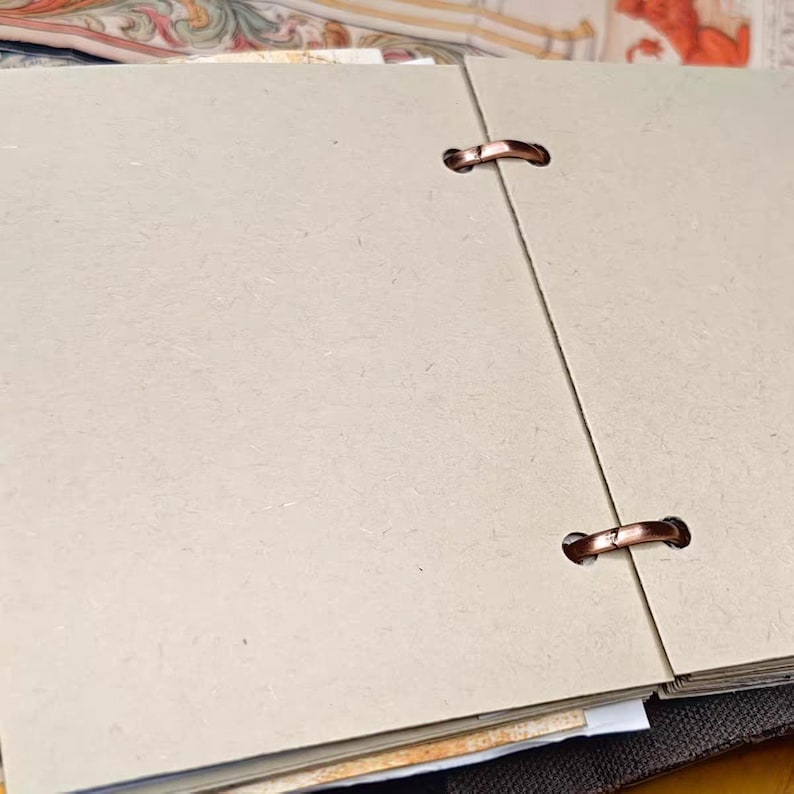 May include: Open journal with blank, light brown pages. The pages are held together with copper-colored metal rings. The journal is partially open, revealing the textured paper and the binding. The background shows a glimpse of a patterned design.