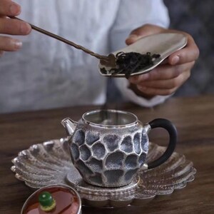 Puede incluir: Un juego de t&eacute; plateado con un dise&ntilde;o texturizado en forma de panal. El juego incluye una tetera, un platillo y una tapa. Una persona vierte hojas de t&eacute; en la tetera con una cuchara. La tetera tiene un asa negra.