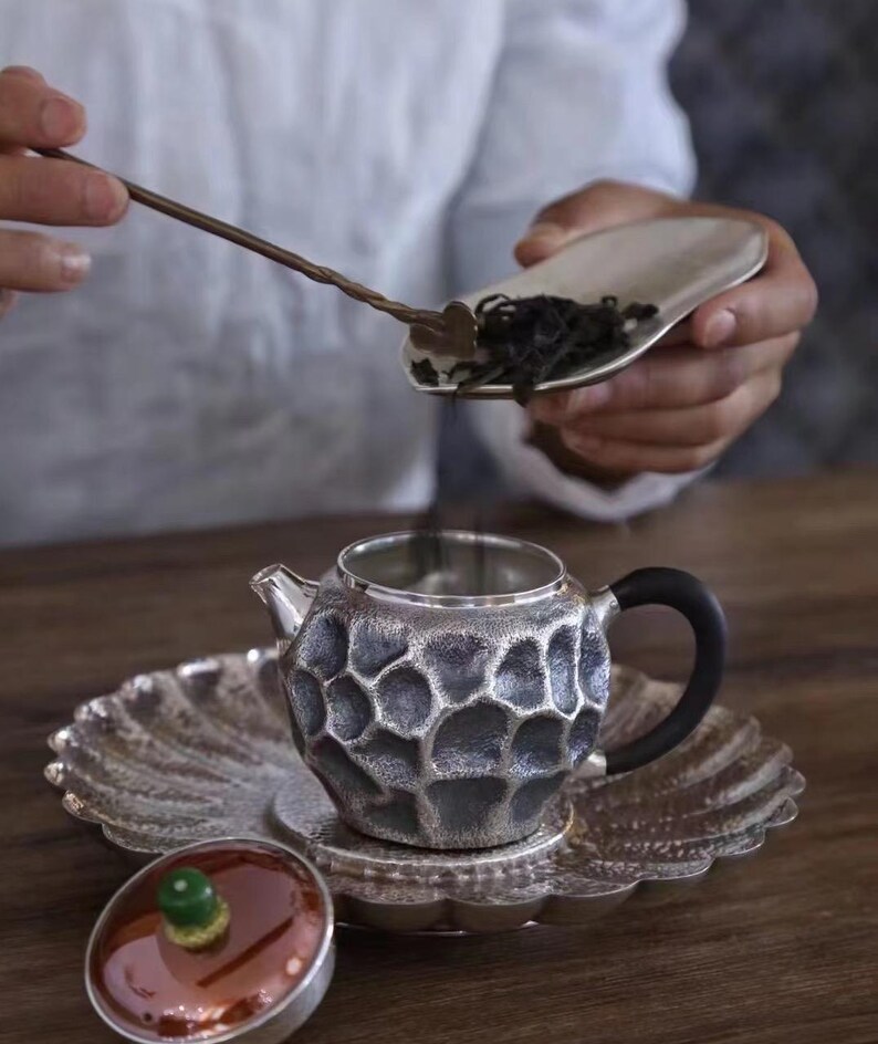 Puede incluir: Un juego de t&eacute; plateado con un dise&ntilde;o texturizado en forma de panal. El juego incluye una tetera, un platillo y una tapa. Una persona vierte hojas de t&eacute; en la tetera con una cuchara. La tetera tiene un asa negra.