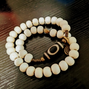 Puede incluir: Pulsera de cuentas de color crema con una cuenta central con estampado en blanco y negro. La pulsera presenta cuentas redondas y detalles metálicos dorados. El diseño se muestra sobre una superficie oscura.