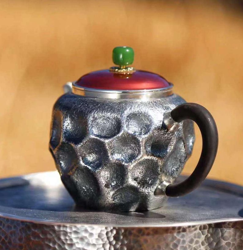 Puede incluir: Una tetera plateada con una superficie texturizada en forma de panal. La tapa es roja con un pomo verde. El asa es de color marr&oacute;n oscuro. La tetera est&aacute; sobre una bandeja plateada con textura martillada.