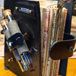 May include: A handcrafted, black leather journal with the text "Philosopher's Stone" on a small metal plate. The journal features a strap with a button closure, holding two glass vials. The pages are aged, with ancient Egyptian hieroglyphs.
