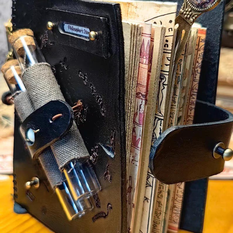 May include: A handcrafted, black leather journal with the text "Philosopher's Stone" on a small metal plate. The journal features a strap with a button closure, holding two glass vials. The pages are aged, with ancient Egyptian hieroglyphs.