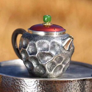 Puede incluir: Una tetera plateada con una superficie texturizada, un asa negra y una tapa roja. La tapa est&aacute; rematada con una piedra verde. La tetera est&aacute; sobre una bandeja plateada. La tetera tiene un dise&ntilde;o &uacute;nico y con hoyuelos.
