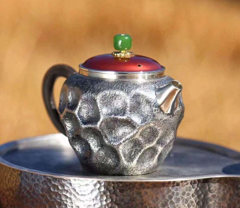 Puede incluir: Una tetera plateada con una superficie texturizada, un asa negra y una tapa roja. La tapa est&aacute; rematada con una piedra verde. La tetera est&aacute; sobre una bandeja plateada. La tetera tiene un dise&ntilde;o &uacute;nico y con hoyuelos.
